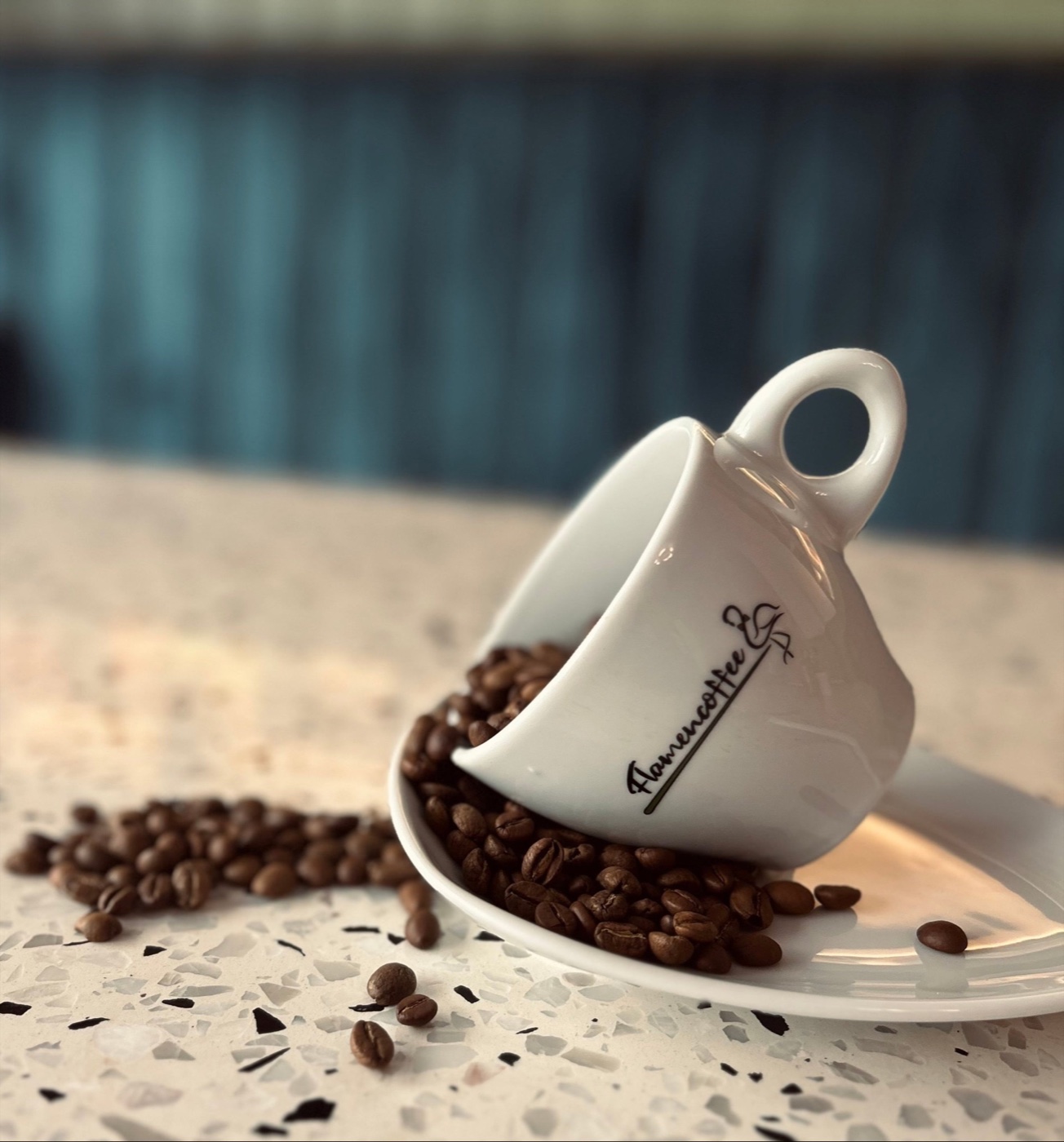 Flamencoffee-Tasse mit herauslaufenden Kaffeebohnen auf dem Terrazzo-Tisch, dahinter die Petrol-Samtbank