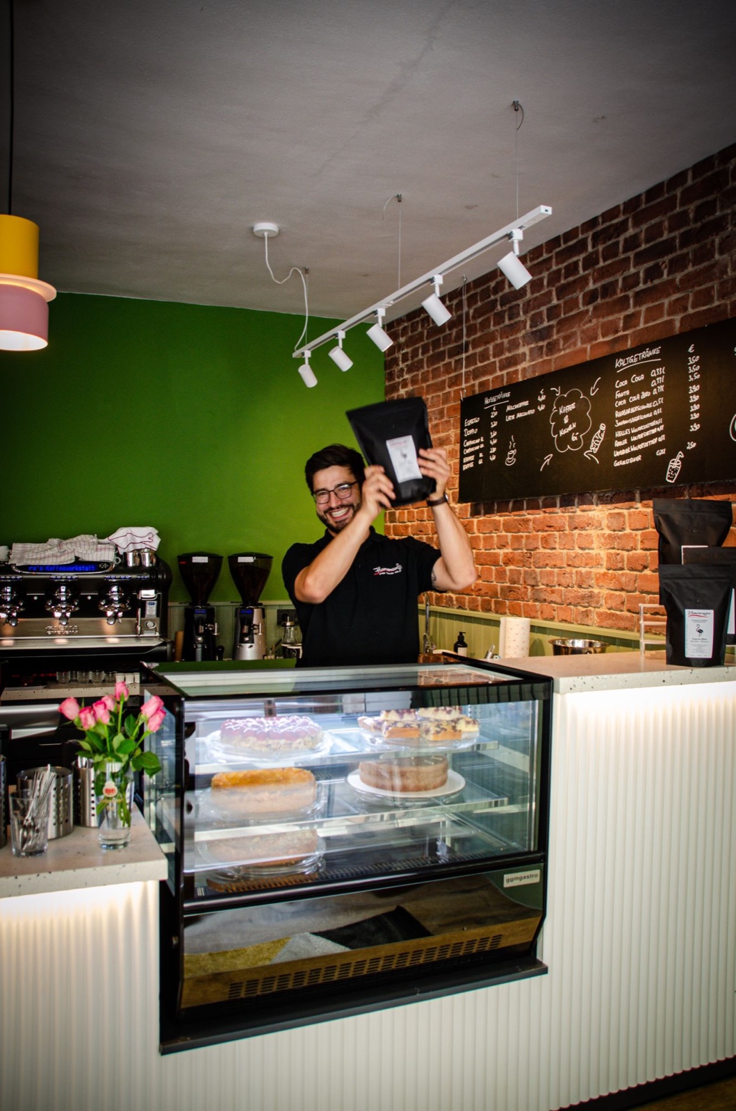 Sandro, der Inhaber von Flamencoffee, strahlt hinter der Kuchentheke und hält eine frisch geröstete Kaffeetüte hoch
